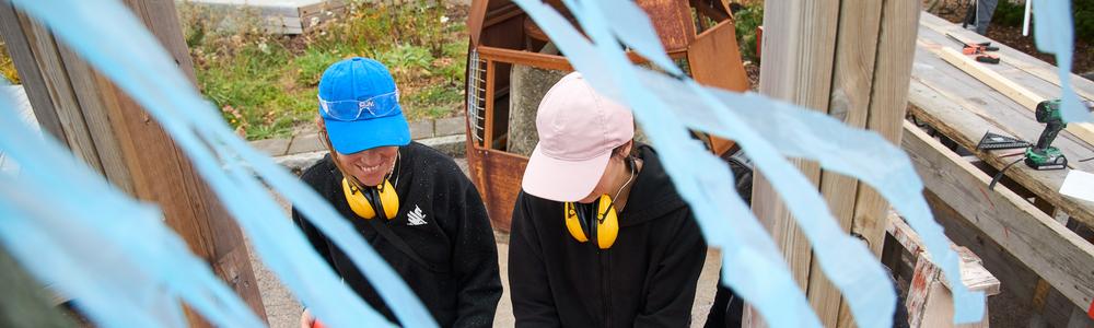 Två personer i keps och hörselkåpor runt nacken bygger en konstruktion med brädor.
